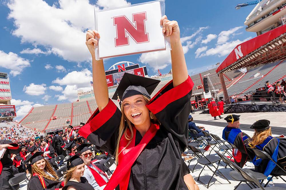 A girl in memorial stadium during commencement holding her diploma up smiling.