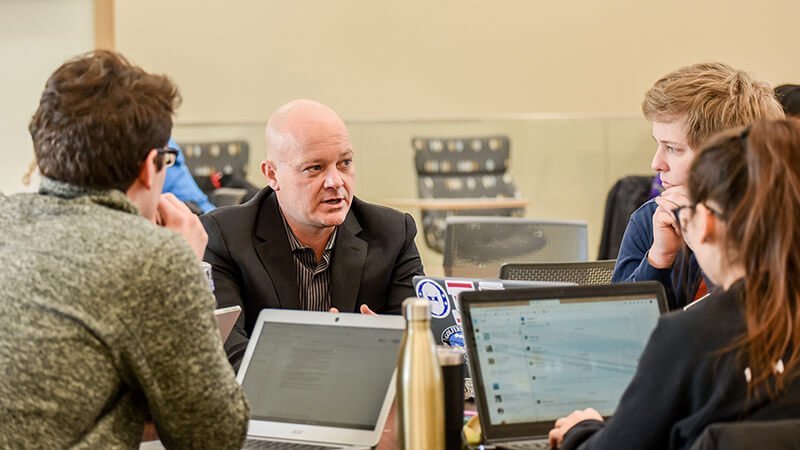 People seated at table with laptops listen to man speaking.