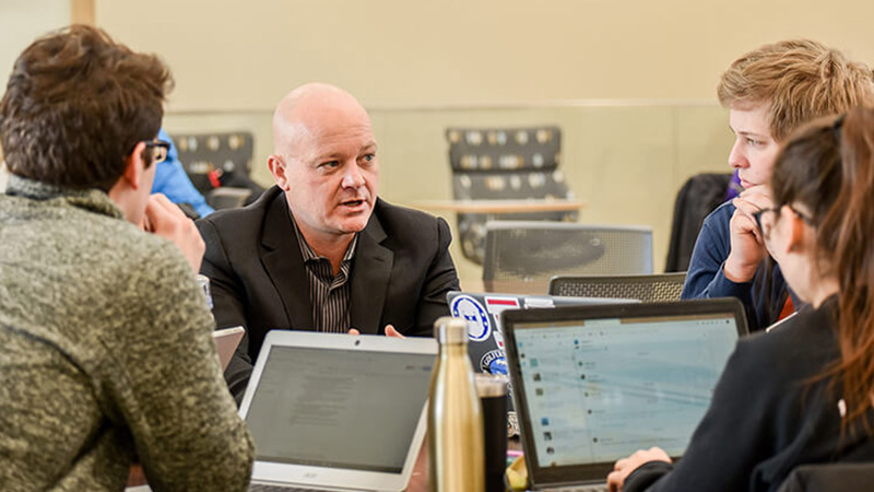 People seated at table with laptops listen to man speaking.