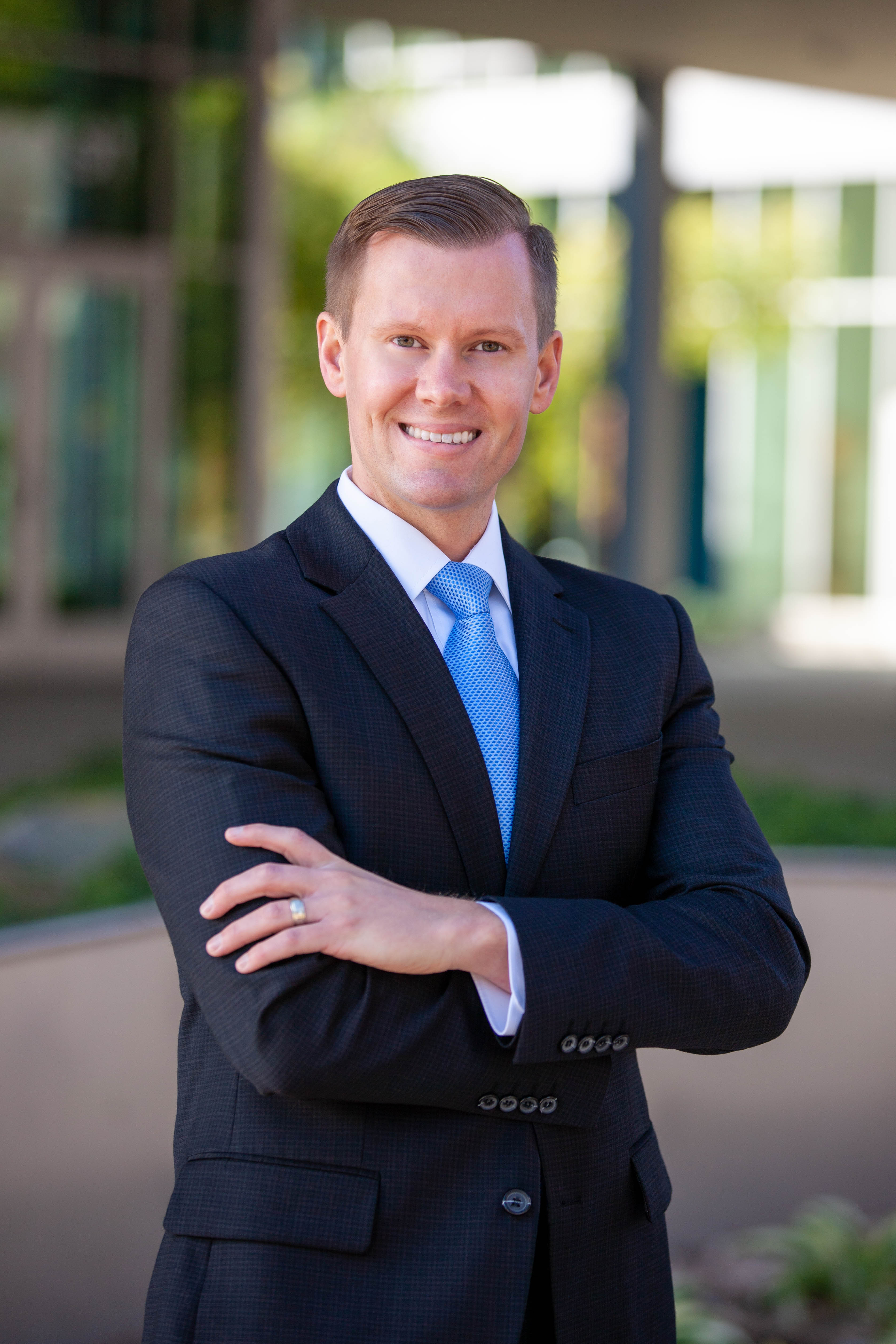 Man in a dark suit and blue tie standing outside with arms crossed, blurred building in background.