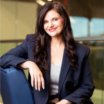 AshLea Allberry; Businesswoman in a navy blazer sitting confidently indoors near a railing.