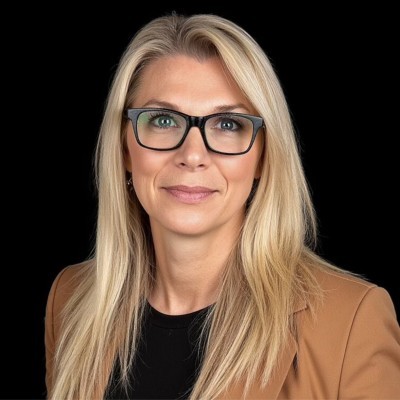 Mandy Dowson; Blonde-haired person wearing a tan blazer over a black shirt, set against a black background.