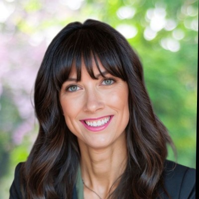 Brett Ebert; A woman with long, wavy dark hair in a blurred outdoor background with greenery and soft pink tones.