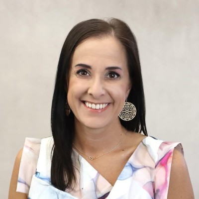 Jess Kinser; Woman in a patterned sleeveless top wearing gold earrings, facing forward with a neutral background.