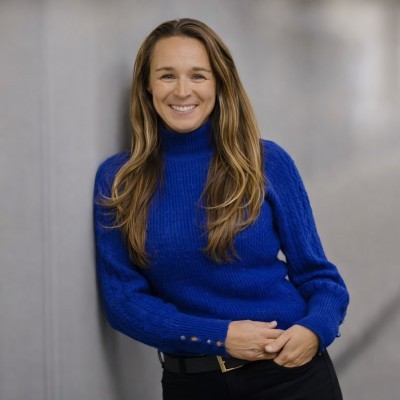 Ashley Krajewski; Woman in a bright blue sweater and black pants leaning against a gray wall indoors.
