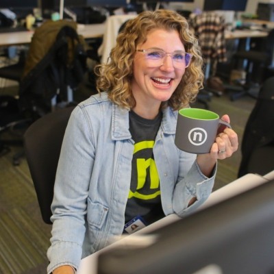 Emily Olinger; Woman seated at a desk, holding a green mug with a circular white logo, indoors in an office setting.
