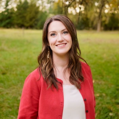Valerie Petersen; woman in a red cardigan standing in a grassy field with trees in the background.