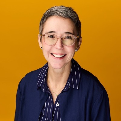 Wendy Townley; Short-haired individual wearing glasses and a blue-striped shirt, set against a solid orange background.