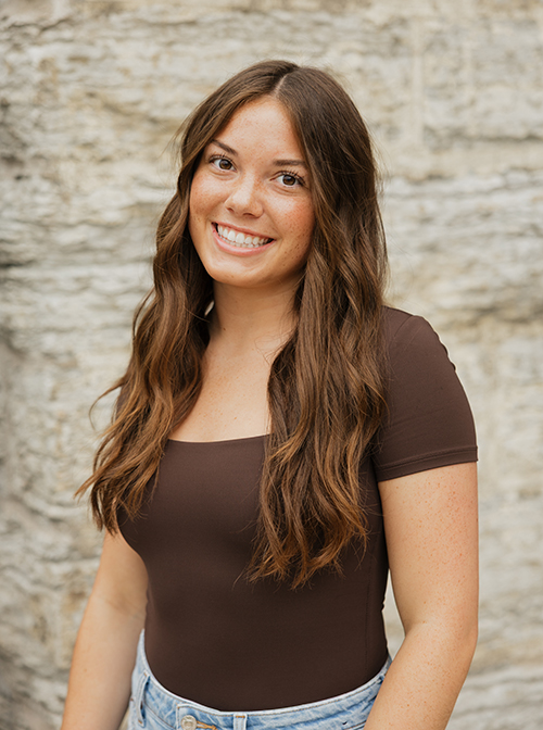 Person with long, wavy brown hair wearing a dark brown top and light blue jeans against a stone wall.