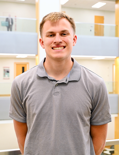 Man in a gray polo stands in a brightly lit indoor space with glass railings and office doors.