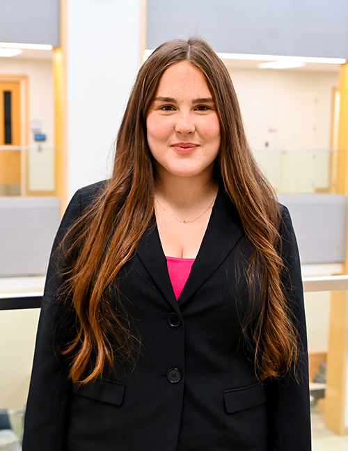 Woman with long brown hair wearing a black blazer and pink shirt, standing indoors.