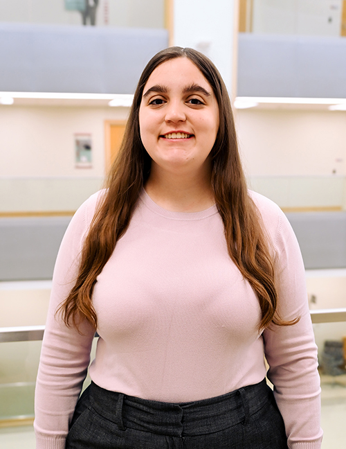 Person standing indoors wearing a pink sweater and dark pants, with long brown hair, blurred face.