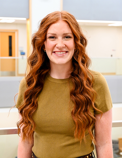 Long curly red hair, wearing a green shirt, standing indoors with blurred background.