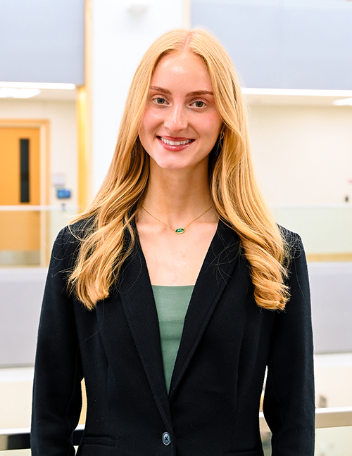 Person with long blonde hair wearing a black blazer and green top in a bright indoor setting.
