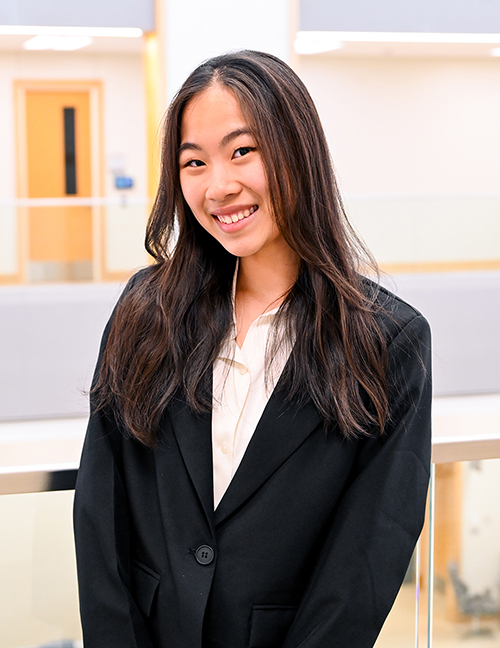 Person wearing a black blazer and cream shirt standing indoors with blurred background.