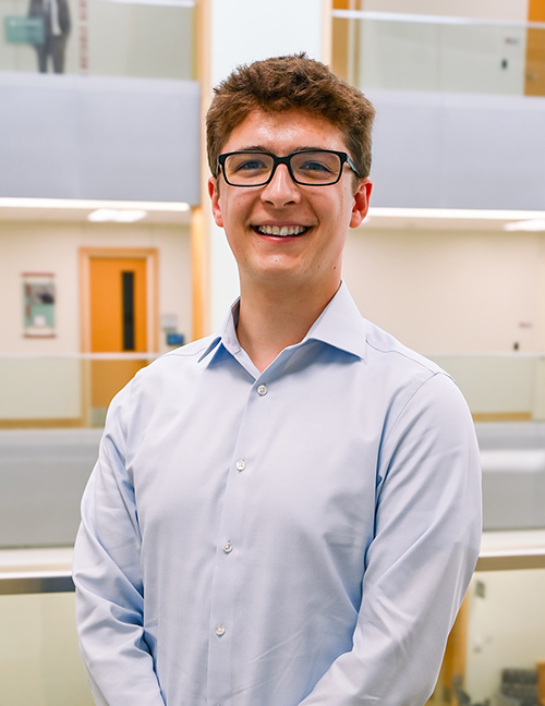 Man in a light blue shirt standing indoors near glass railings and walls.