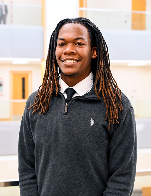Person with long braided hair wearing a dark sweater and tie in a modern indoor setting.