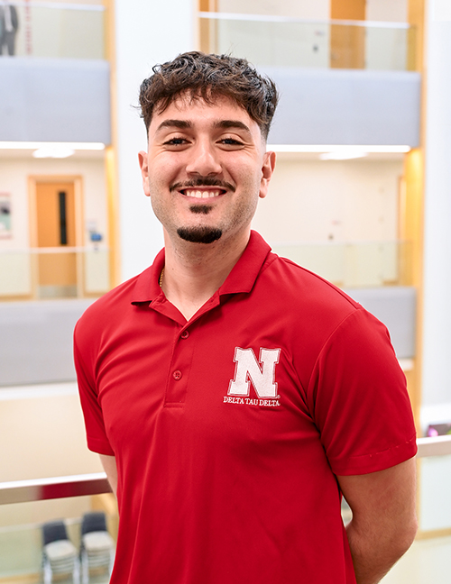 Man in red polo with Delta Tau Delta logo, standing indoors in front of a modern, multi-level building.