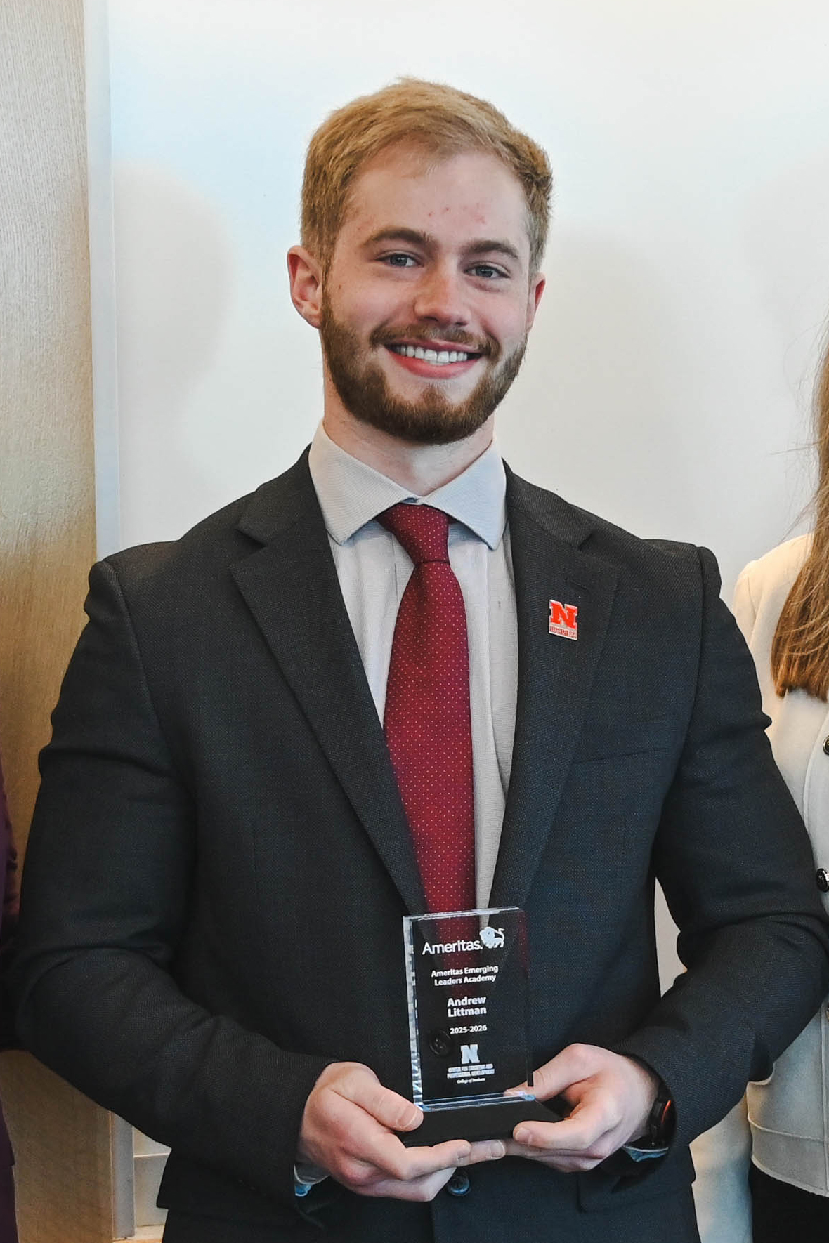 Man in a suit holding a trophy, standing indoors against a white wall.