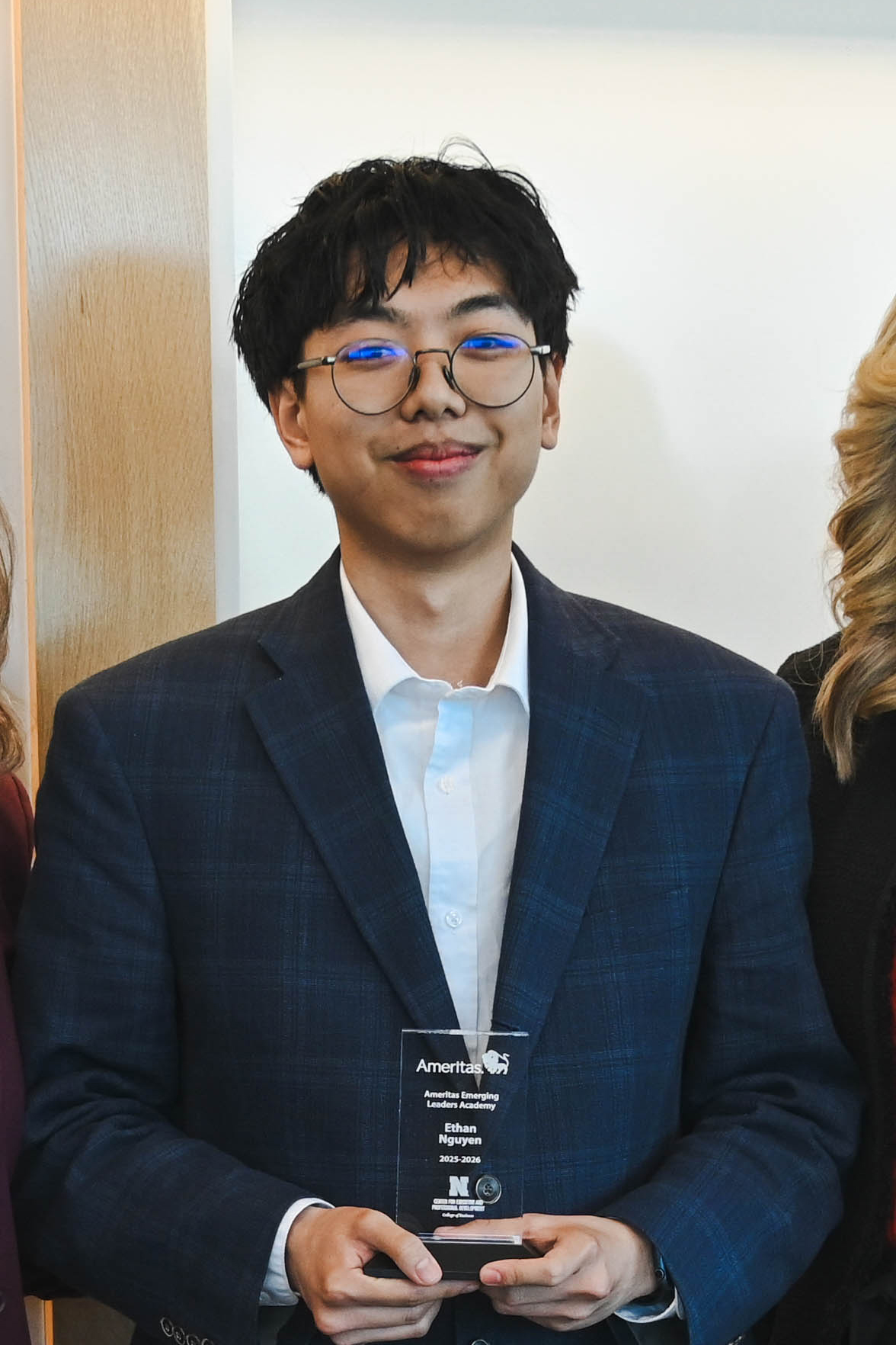 Man in a blue blazer holding a glass award, standing indoors with others partially visible.