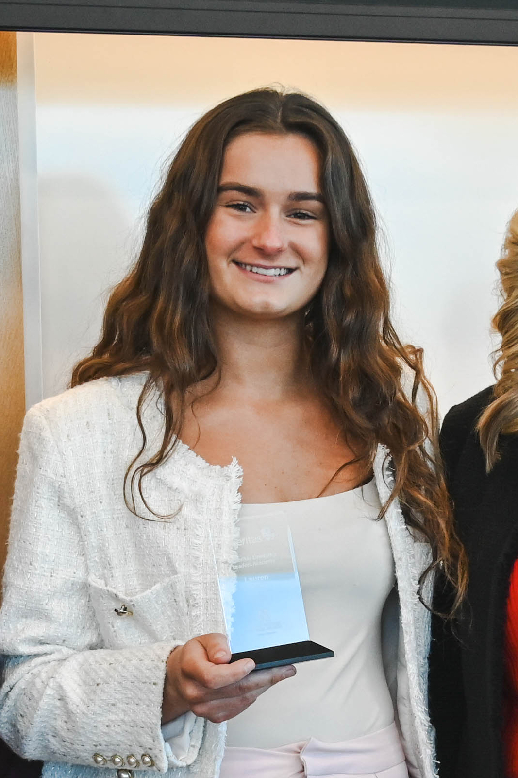 Woman holding a glass award, wearing a white outfit, standing indoors.