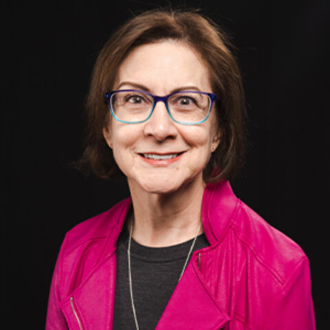 Person wearing a pink jacket and gray top against a black background. Face blurred.