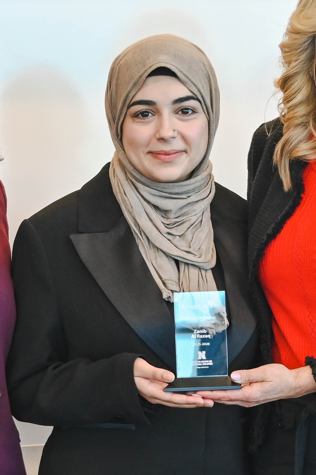 Person in a beige hijab and black suit holding a glass award, standing beside others.