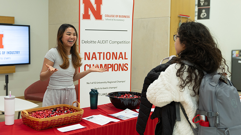 Booth display with banner promoting college competition, person speaks to visitor with backpack.
