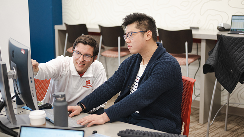 Two people seated at desks, collaborating while looking at a computer screen in a study space.