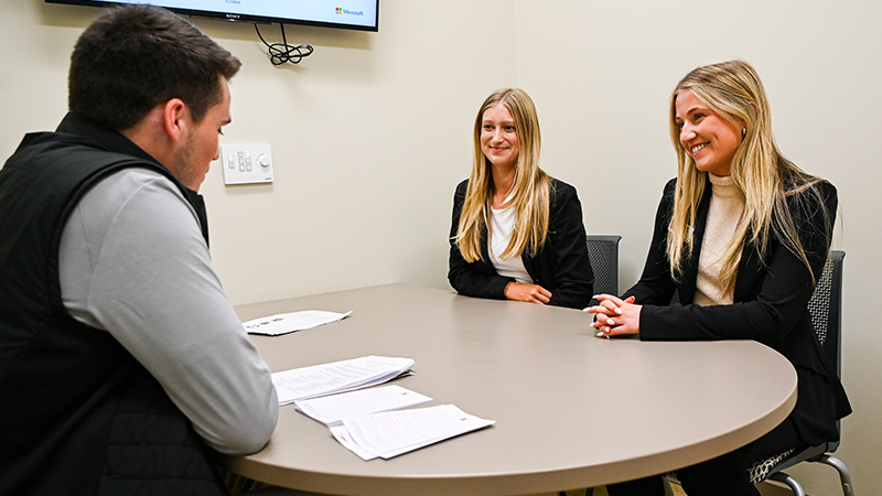 Three people in a meeting, seated at a round table with documents placed on it.