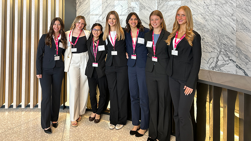 Seven women in business attire with pink lanyards stand together in a modern, marble-accented space.