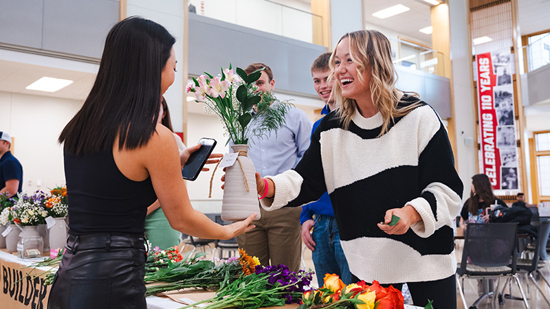 Woman handing vase of flowers to another in a bright indoor market setting.