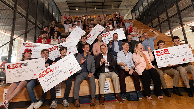 Group seated on tiered steps, holding large ceremonial checks and smiling in a bright indoor space.