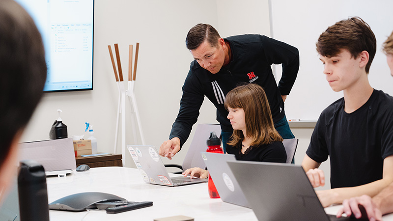 Group working at laptops in a classroom, instructor helping a seated student. 