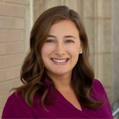 Riley Illg; Smiling woman with wavy brown hair, wearing a purple blouse, standing near a brick wall indoors.