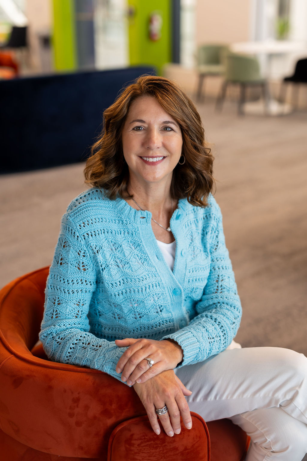 DeeAnn Wenger; Woman seated on an orange chair, wearing a blue cardigan, smiling in a modern indoor setting.