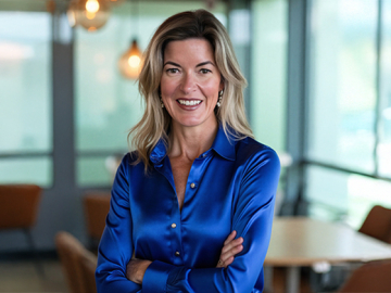 Jennifer Lea: Smiling woman in a blue satin blouse standing with arms crossed in a modern, softly lit room.