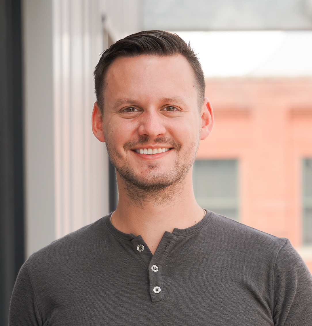 Brett Byman; Man smiling in a gray shirt, standing outdoors near a building.
