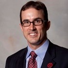 Man in a suit and tie smiling, wearing glasses against a neutral background.
