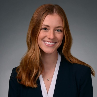 Camille Sippel; Smiling woman with long red hair wearing a dark blazer and white blouse against a gray background.
