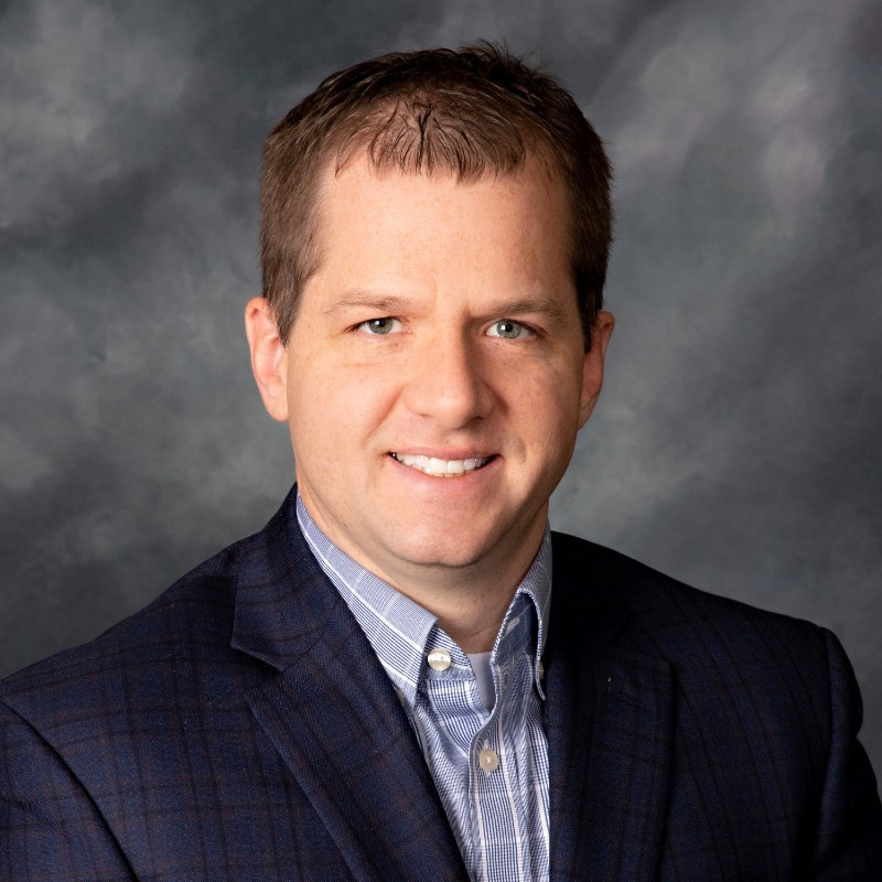 Smiling man in business attire, with a dark plaid blazer and light blue shirt, set against a cloudy backdrop.
