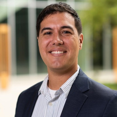 Smiling man in a suit and striped shirt, standing outdoors with blurred building and greenery behind.