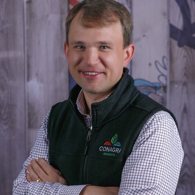 Smiling man in a dark vest and checked shirt with crossed arms against a wooden background.