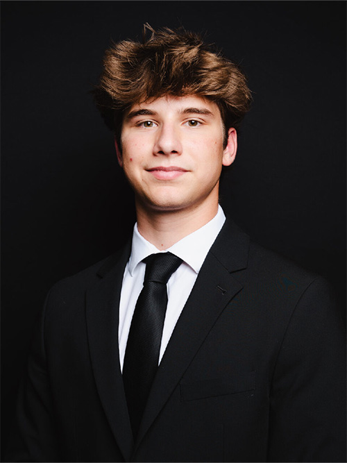 Young man in a black suit and tie against a dark background, with a neutral expression.