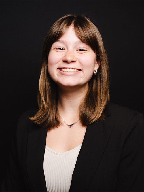 Smiling woman with shoulder-length brown hair, wearing a black blazer over a light top, on a black background.