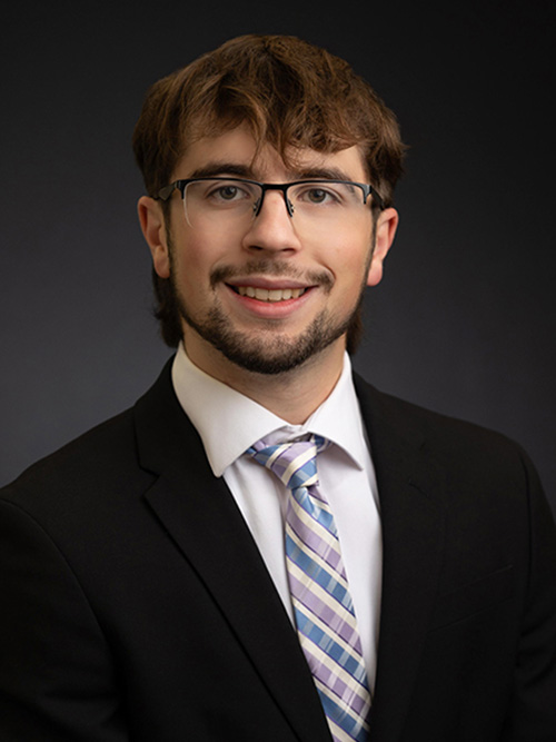 Man wearing glasses, a dark suit, white shirt, and striped tie, smiling in front of a dark background.