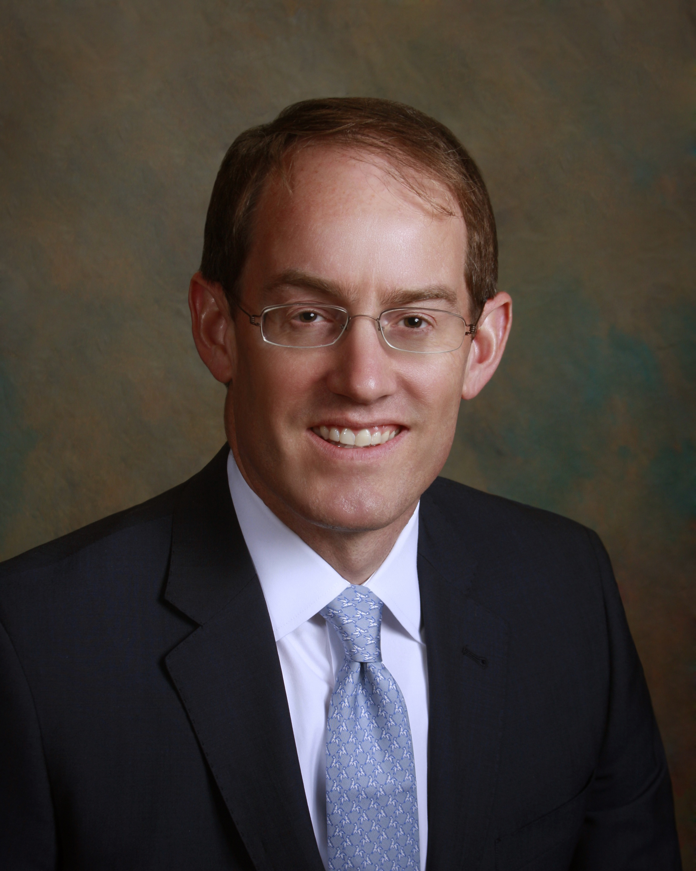 Man in a suit with a blue tie, smiling, against a textured brown and green backdrop.