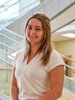 Smiling woman in a white shirt standing near glass stair railings indoors.