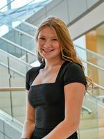 Smiling woman with long hair in a black dress, standing near a modern glass staircase indoors.