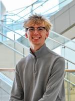 Smiling young man in glasses and a gray sweater standing indoors with stairs in the background.